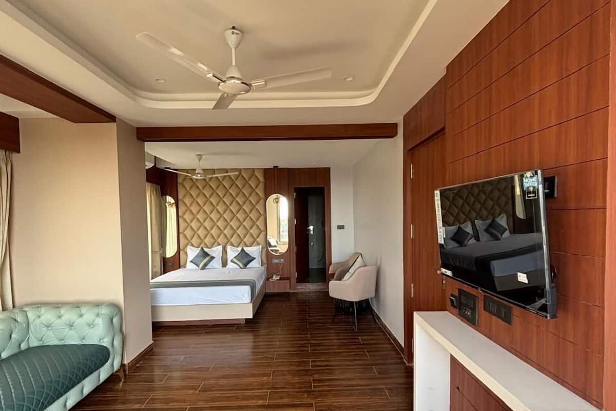 A spacious hotel room with a large bed, a sofa, a TV mounted on the wall, and a ceiling fan.