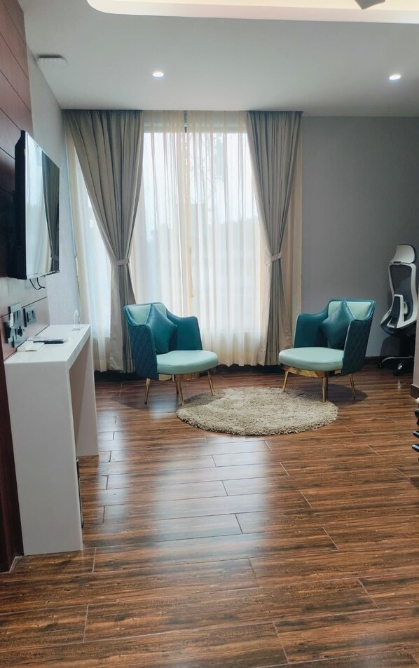A modern living room with a wooden floor, two teal chairs, a white desk, a large window with curtains, and a ceiling light. - HOTEL BIVA (Silchar)