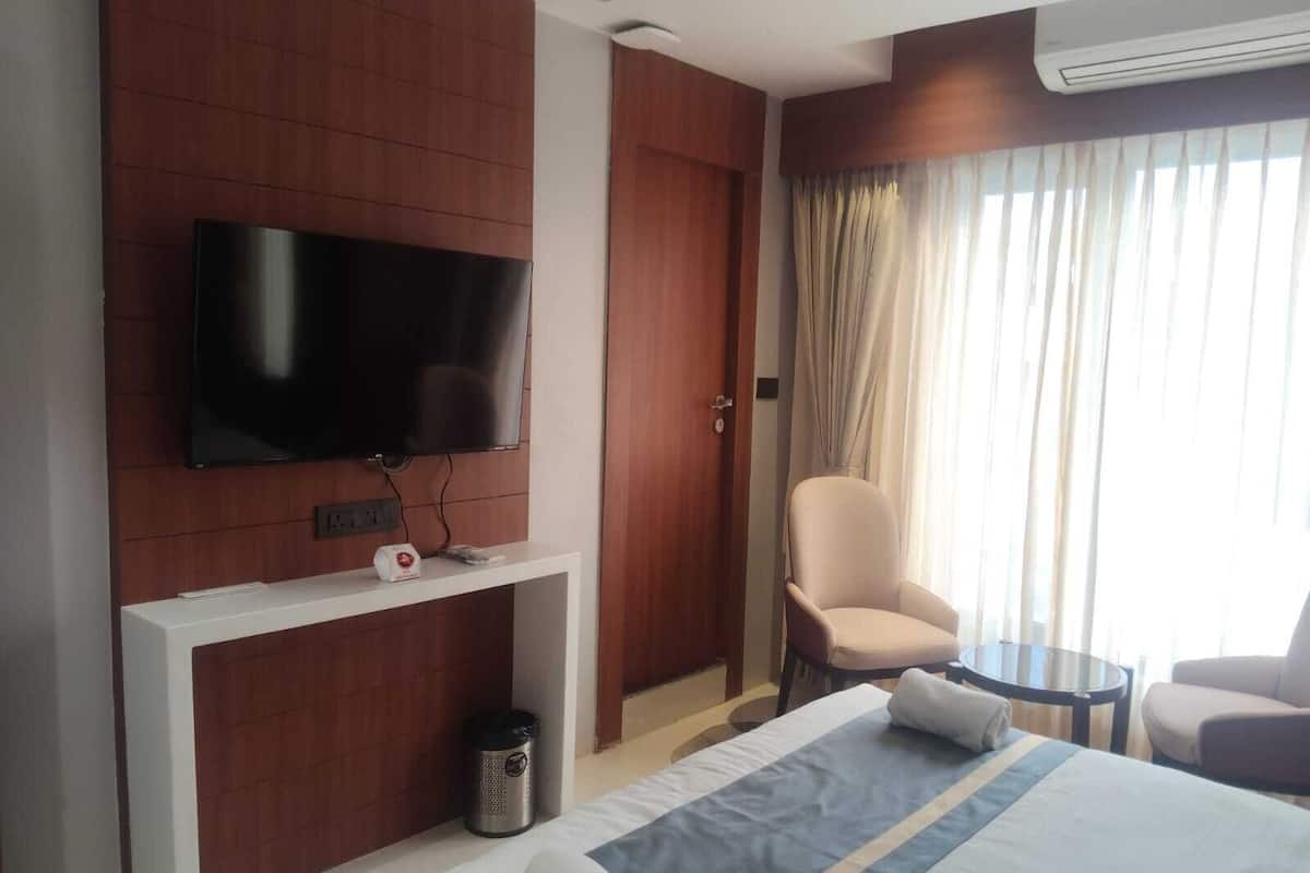 A hotel room with a flat-screen TV mounted on a wooden panel, a bed with a blue and white striped bedspread, a chair, a small round table, and a window with curtains.
