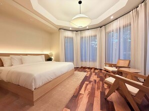 Une chambre moderne avec un grand lit, un plancher en bois, un coin salon avec une table et de grandes fenêtres ornées de rideaux légers.