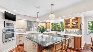 A modern kitchen with a central island, stainless steel appliances, wooden cabinets, and a large window providing a view of greenery. - Deer Run Lodge/Indoor Pool/Basketball/Hot Tub (Terre Haute)