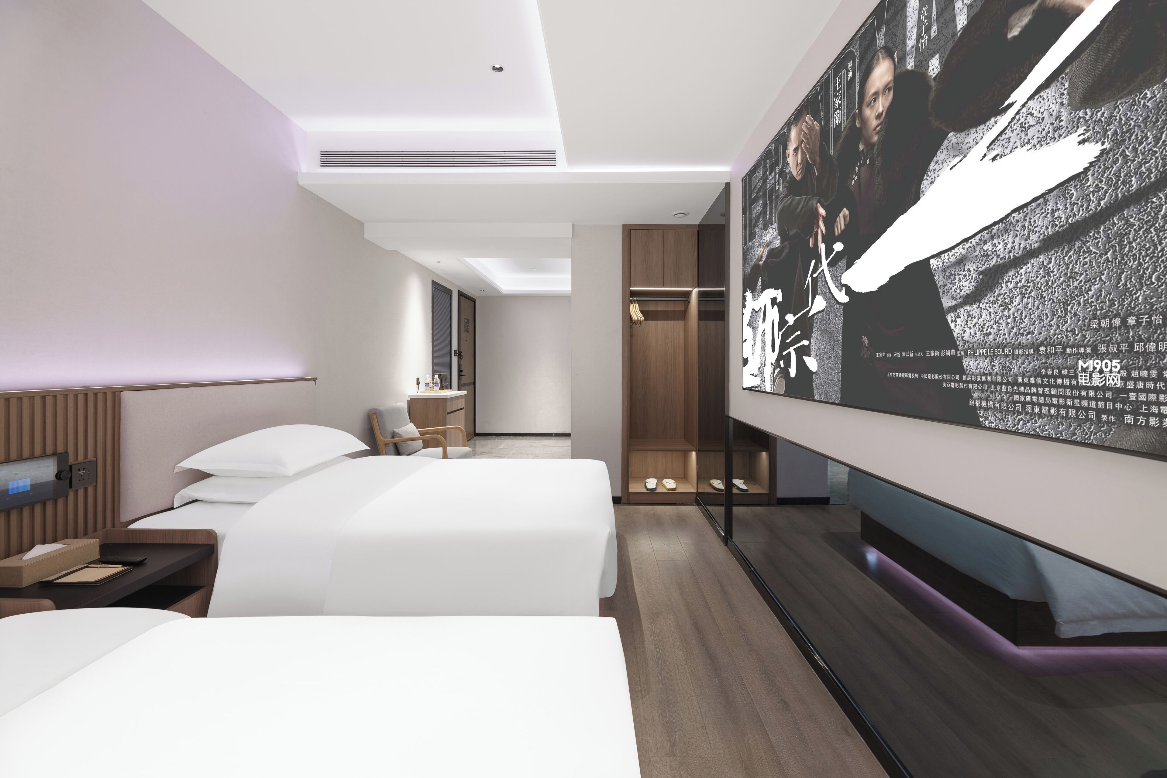 A modern hotel room with two beds, a desk, a wardrobe, and a large TV screen displaying a movie.