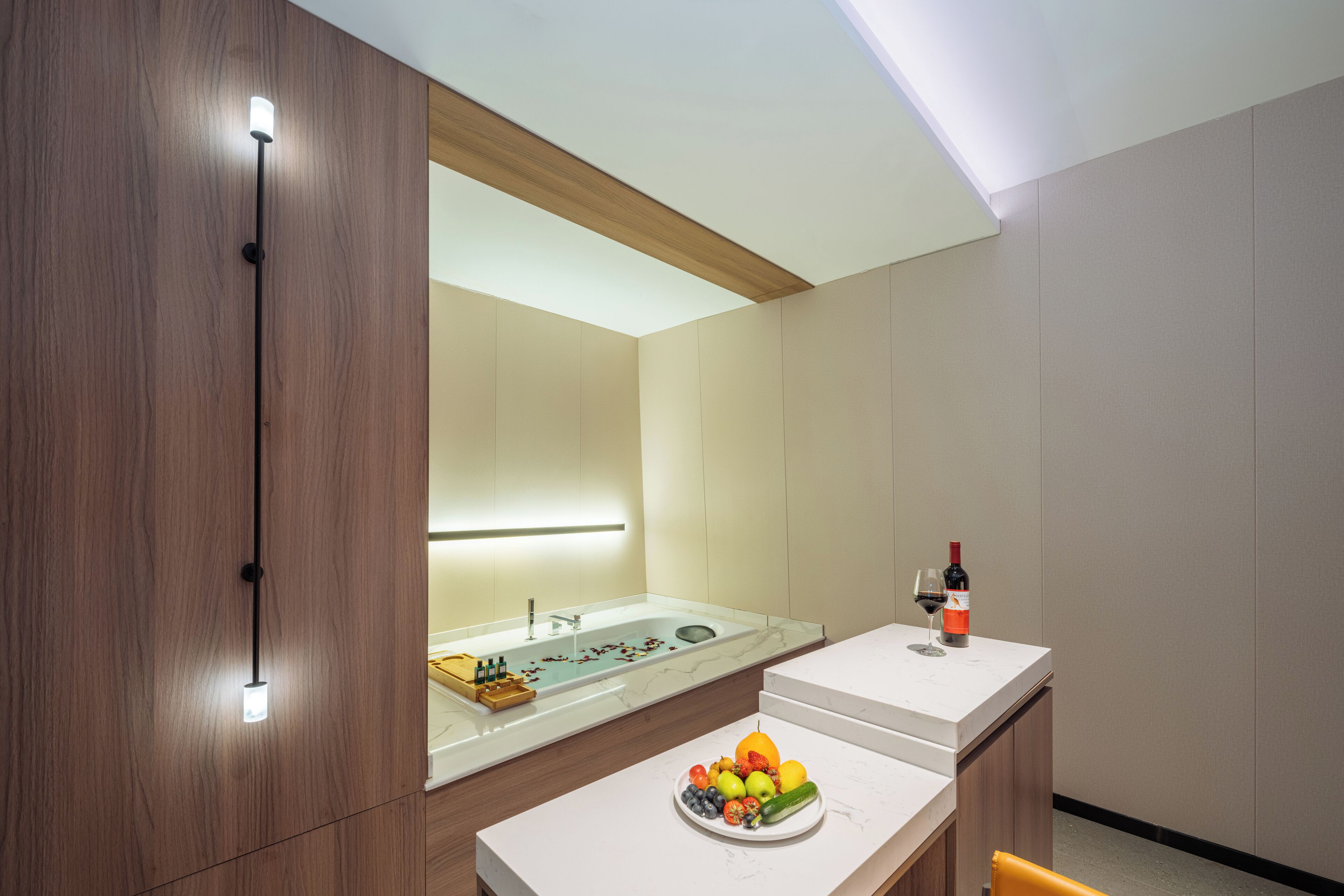 A modern bathroom with a wooden vanity, a large mirror, a sink with a marble countertop, and a fruit bowl with a bottle of wine on the counter.