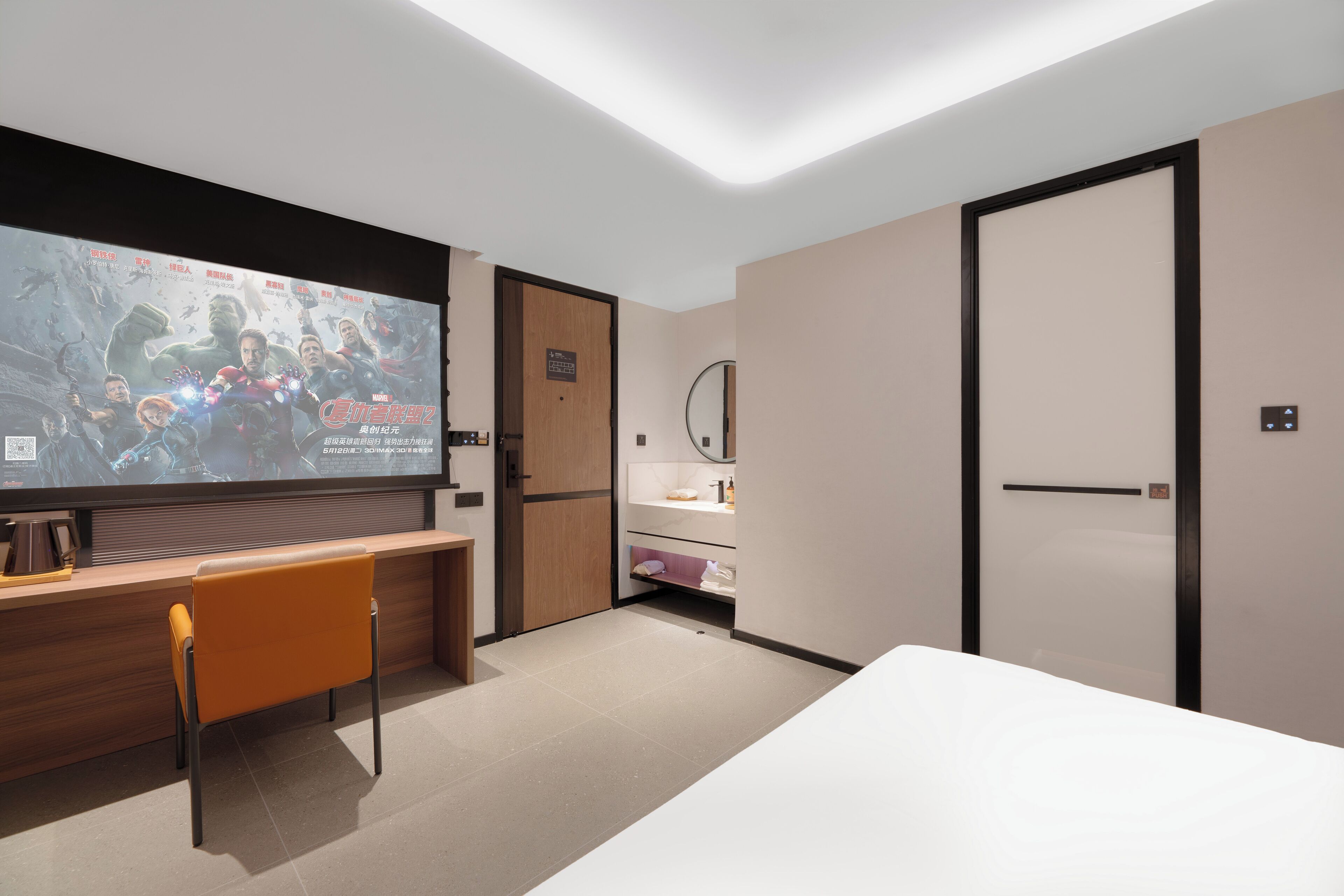 A hotel room with a large flat-screen TV mounted on the wall, a wooden desk, a chair, a bed, a bathroom with a mirror, and a door.