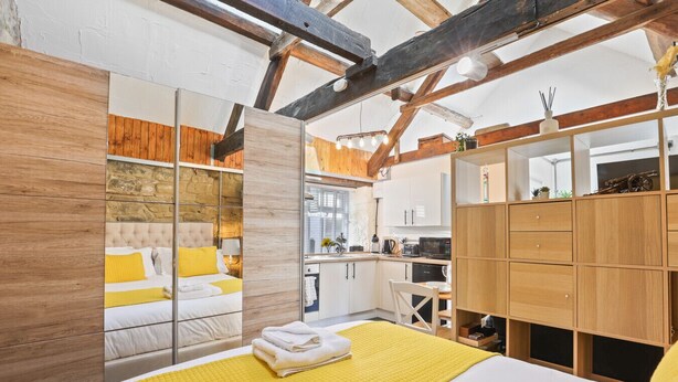 A modern bedroom with a large bed, a wooden wardrobe, and a kitchen area visible through an open door.