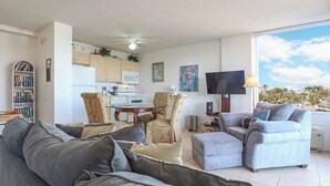 A living room with a grey sofa, a dining area with a table and chairs, a kitchen with a microwave, and a TV mounted on the wall.