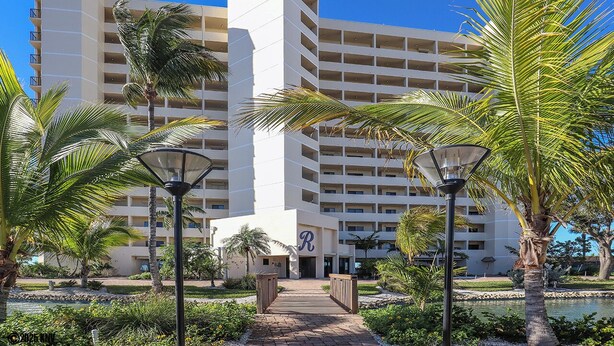 A multi-story hotel with a central entrance, surrounded by palm trees and a pathway leading to the entrance. - Riviera Beach Club 1102 - 2B/2B Dream Vacation Condo Amazing Gulf Views! (Fort Myers Beach)