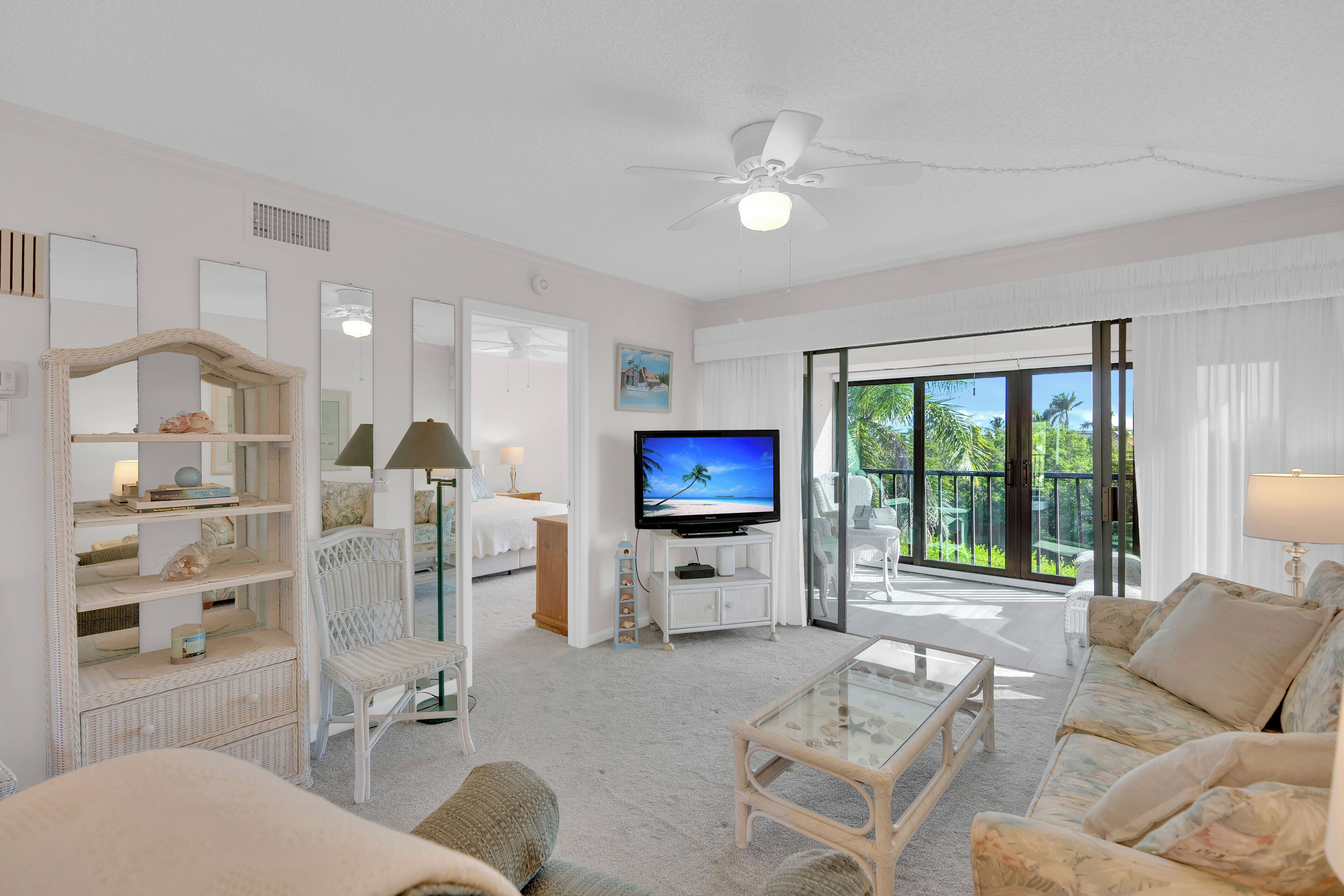 A spacious living room with a flat-screen TV, wicker furniture, a ceiling fan, and a large sliding glass door leading to an outdoor area.