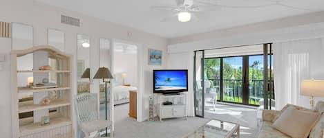 A spacious living room with a flat-screen TV, wicker furniture, a ceiling fan, and a large sliding glass door leading to an outdoor area.