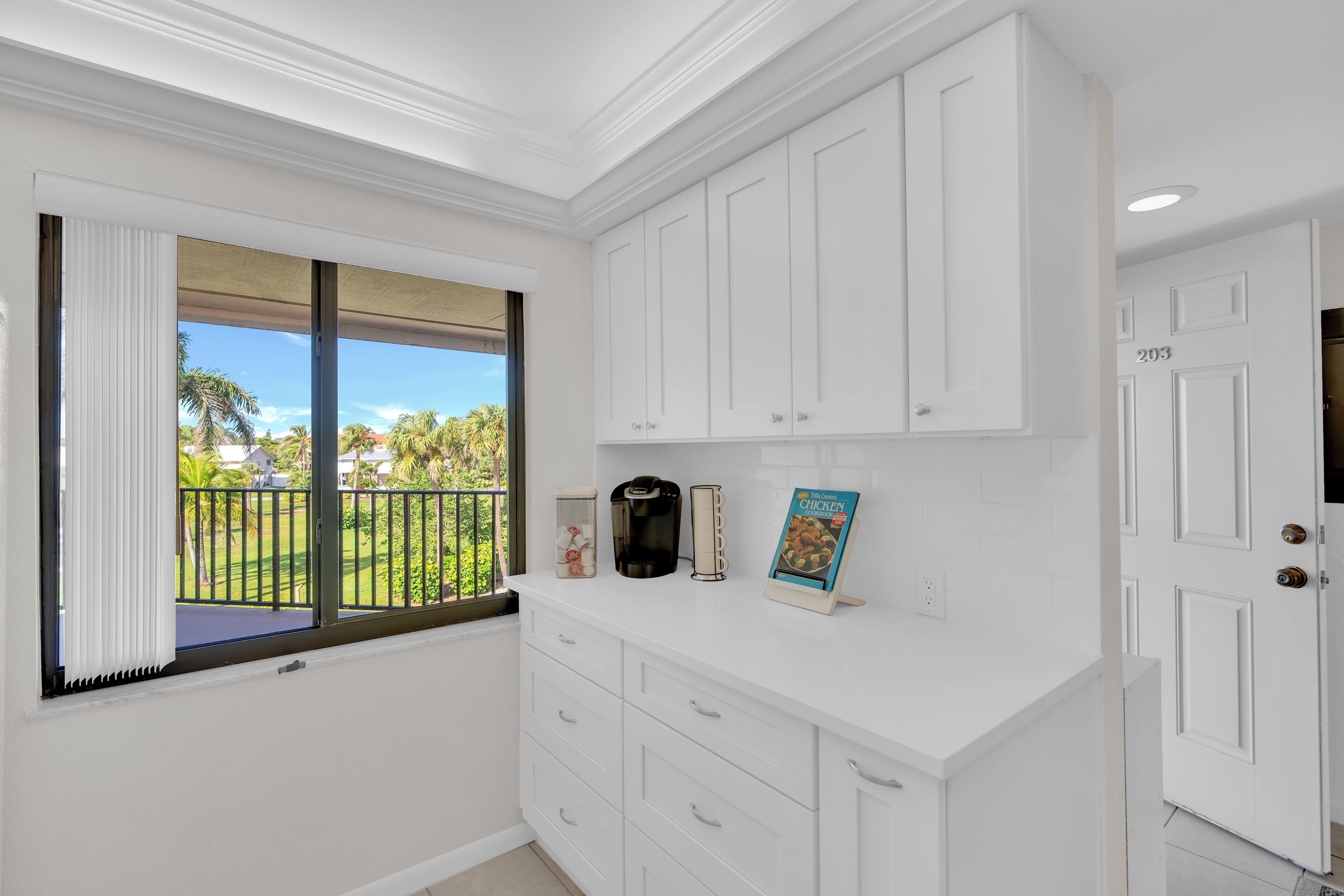 A kitchen with white cabinetry, a coffee maker, a cookbook, and a view of a green lawn through a window.