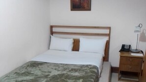 Uma cama bem arrumada com um cobre-leito estampado, uma mesa de cabeceira com abajur e um pequeno quadro na parede.