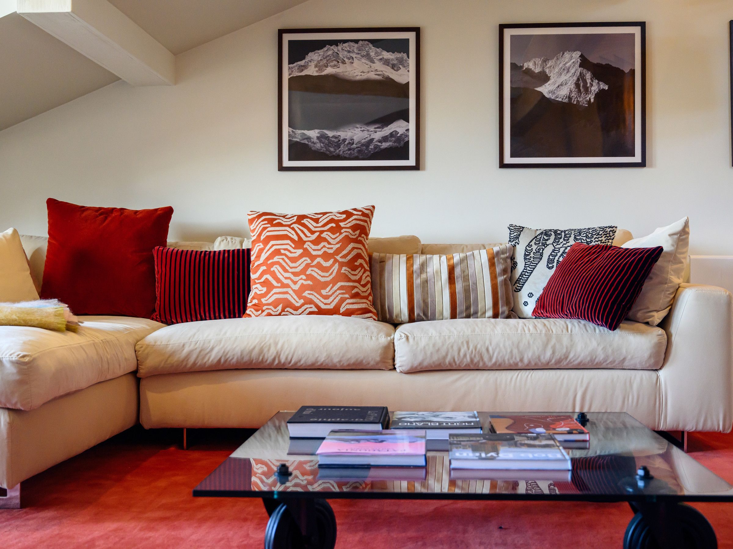 A living room with a beige sectional sofa, red and patterned cushions, a glass coffee table with books, and two framed mountain landscape pictures on the wall.