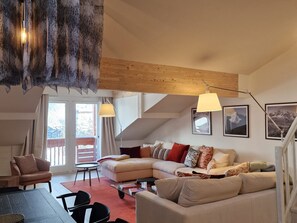 A spacious living room with a sectional sofa, a small round table, and a coffee table. There is a balcony with a view of the outdoors.