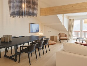 A modern living room with a dark wooden dining table, black chairs, a white sofa, and a wooden ceiling beam.