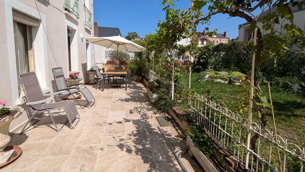 A patio with a table and chairs, a white umbrella, and a garden with a fence.