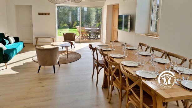 Un coin repas avec une table en bois dressée pour six personnes, entourée de chaises en bois, et une grande fenêtre donnant sur un jardin.
