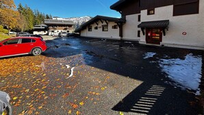 A parking area with a red car, a building with a white facade and dark trim, and a mountain backdrop.