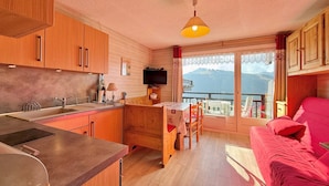 A cozy kitchen with wooden cabinets, a sink, and a dining area with a table and chairs. A red sofa is visible in the foreground, and a balcony with a view of mountains is seen through the window.