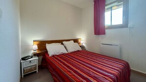 Une petite chambre propre, avec un lit simple, une table de chevet, une lampe et une fenêtre avec des rideaux.