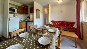 Une cuisine compacte équipée d’un four à micro-ondes, d’un réfrigérateur et d’un évier. Une table à manger avec une nappe à pois et des chaises est dressée pour le repas. Un canapé rouge à motifs en forme de cœur est placé contre le mur.