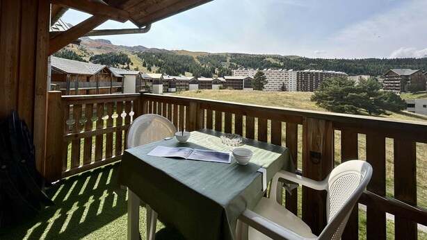 Une terrasse en bois avec une table et des chaises, offrant une vue sur un paysage montagneux parsemé de bâtiments.
