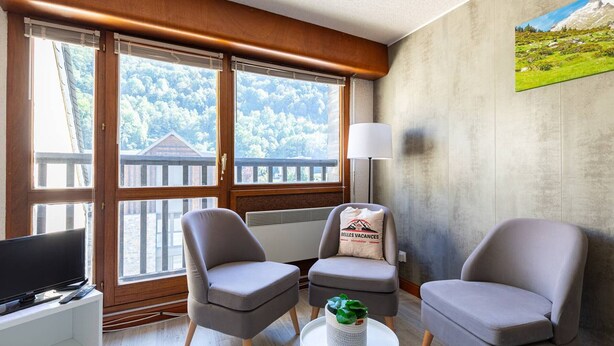 A modern living room with a large window, two grey armchairs, a small table with a plant, a TV, and a lamp.