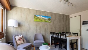 A modern living room with a sofa, armchair, dining table, and chairs. A lamp and a painting are on the wall.