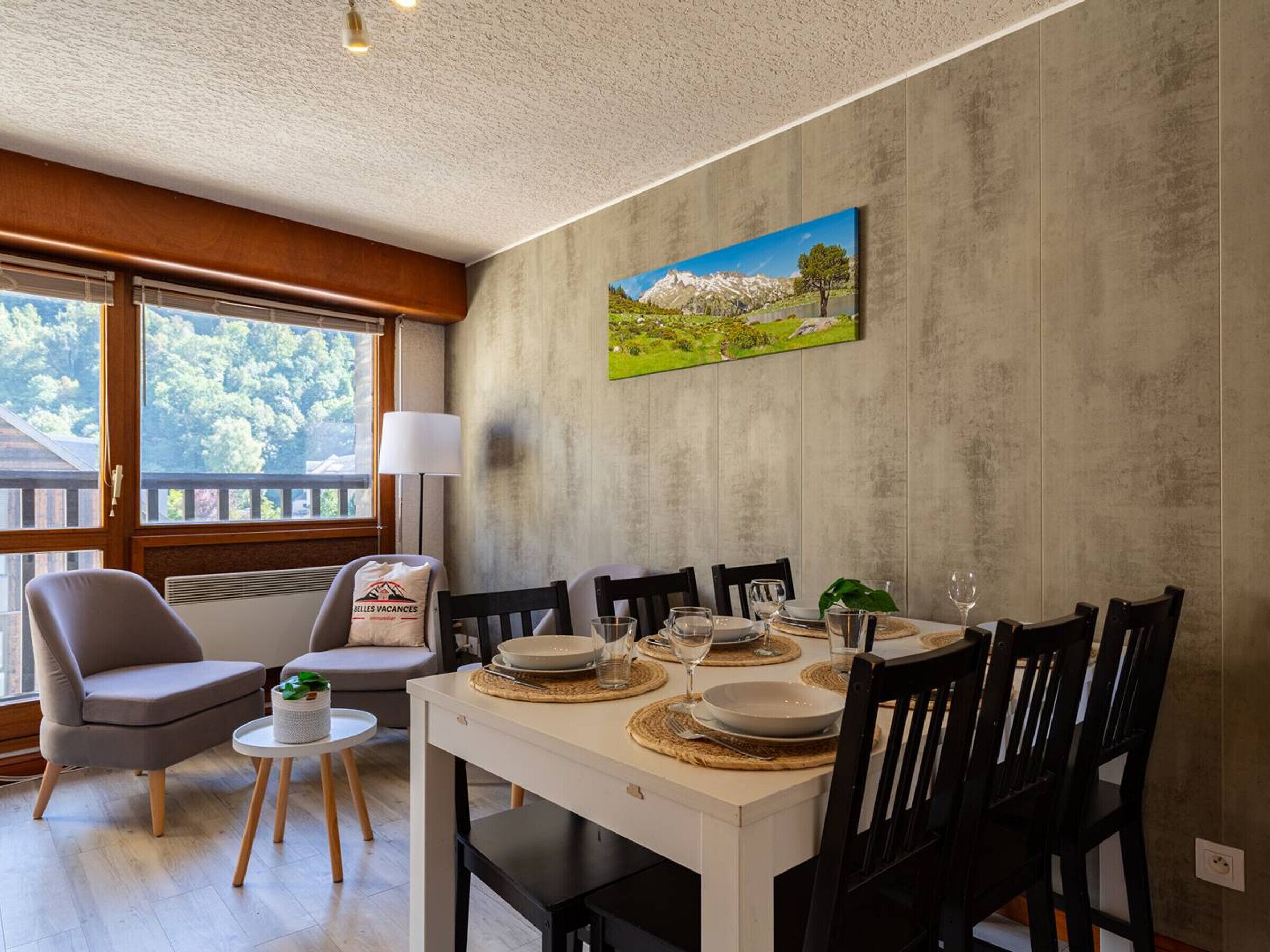 Un comedor con una mesa blanca dispuesta para cuatro personas, rodeada de sillas negras. Un cuadro de un paisaje cuelga en la pared.