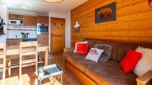 Een gezellig chaletinterieur met houten details, een kitchenette, een eethoek en een woonkamer met een bank en kussens.
