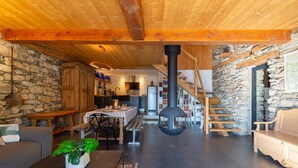 Un interior acogedor con pared de piedra, vigas de madera en el techo, una chimenea central, un comedor y una escalera que conduce a un nivel superior.