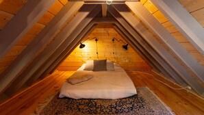 Un dormitorio acogedor en el ático, con techo inclinado, vigas de madera, una cama con almohadas, una alfombra y dos lámparas fijadas a la pared.