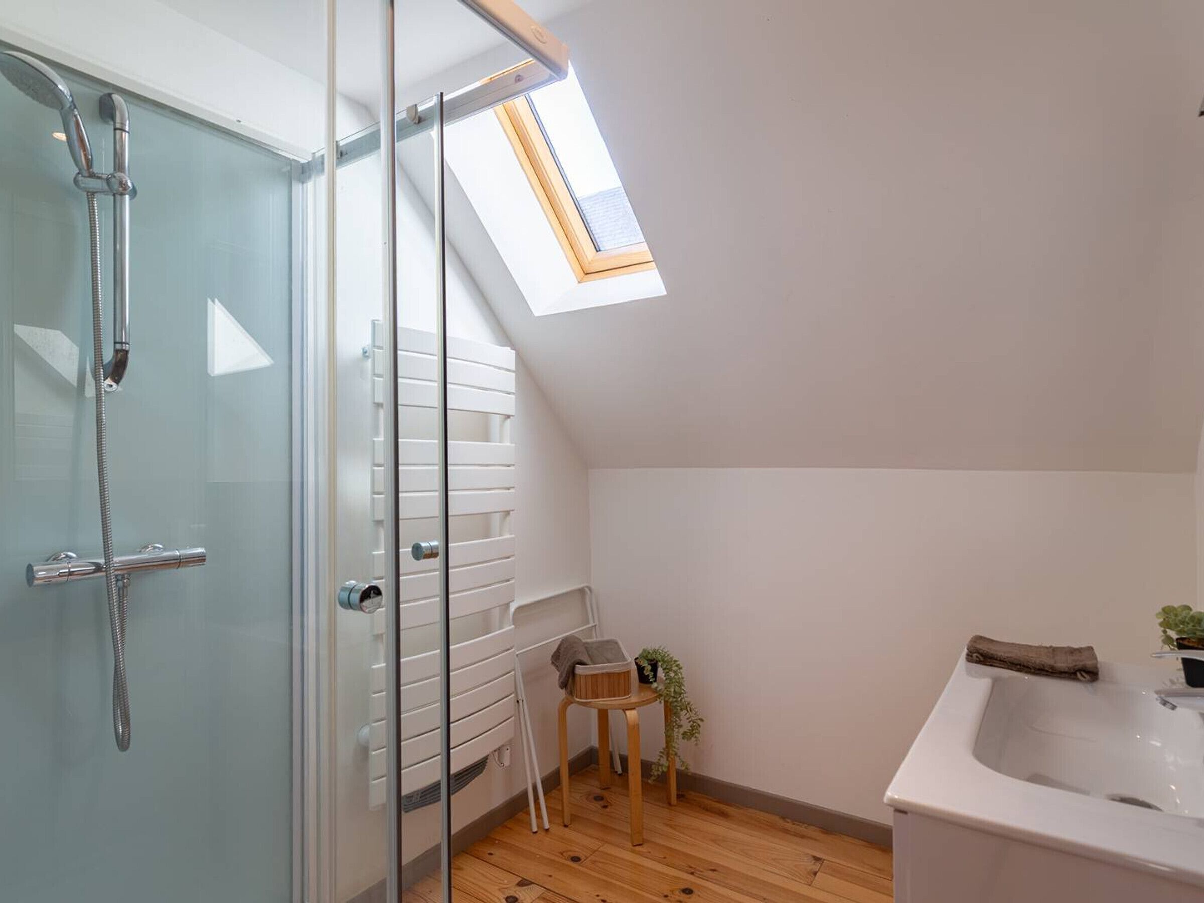 A modern bathroom with a glass shower enclosure, a skylight, a white sink, and a small wooden stool.