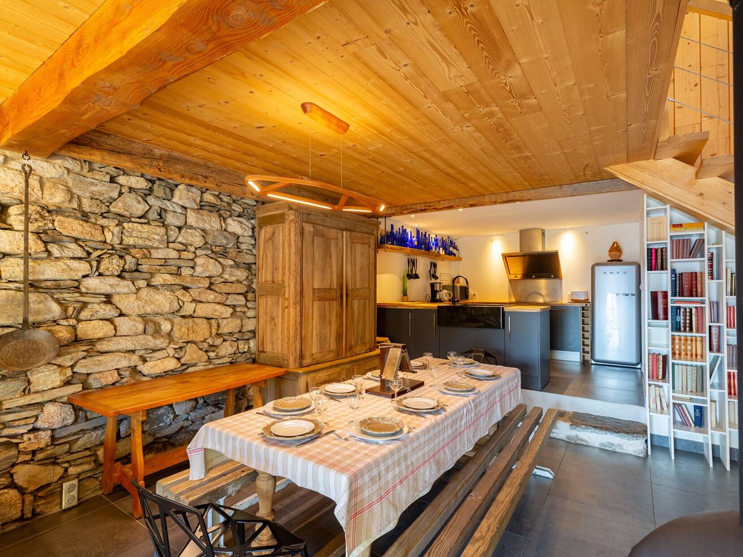 A cozy dining area with a stone wall, wooden table, and benches. A kitchen with modern appliances and a bookshelf is visible in the background.