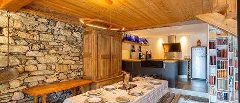 A cozy dining area with a stone wall, wooden table, and benches. A kitchen with modern appliances and a bookshelf is visible in the background.
