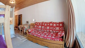 Ein gemütlicher Raum mit Holzboden, einem Sofa aus Holz mit gemustertem Bezug, einem Tisch mit Stühlen und einem Fenster mit Vorhängen.