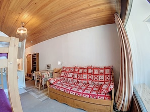Ein gemütlicher Raum mit Holzboden, einem Sofa aus Holz mit gemustertem Bezug, einem Tisch mit Stühlen und einem Fenster mit Vorhängen.