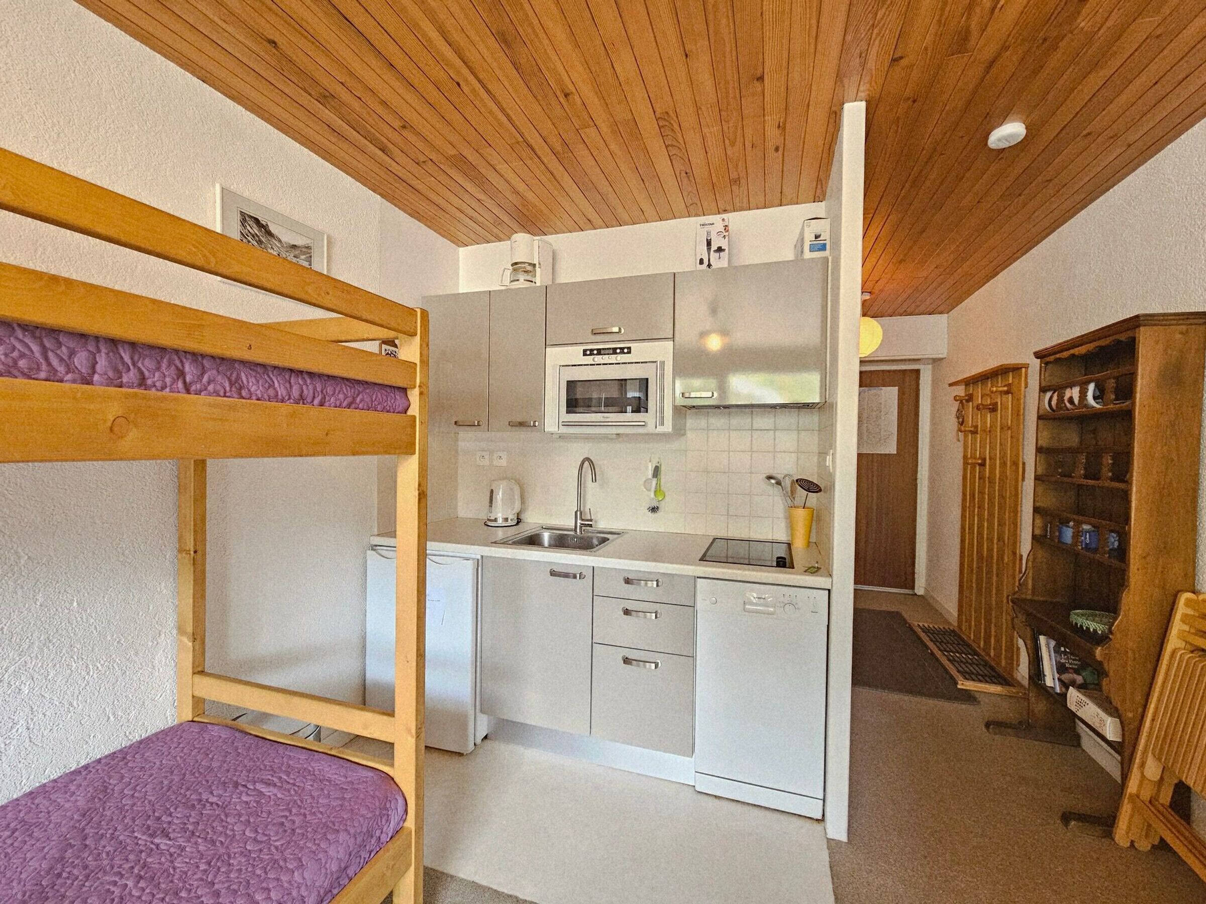 Cocina compacta con microondas, pileta y alacenas, junto a una cama litera con colchón morado.