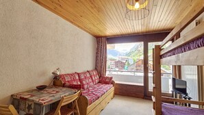 Ein gemütliches Zimmer mit Holzbalkendecke, einem Sofa mit gemusterten Kissen, einem Tisch mit Schale und Blick auf ein Bergdorf durch ein Fenster.
