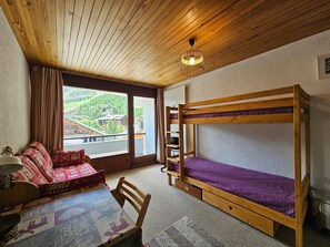 Ein Zimmer mit einem Etagenbett, einer Schlafcouch, einem Tisch und einem Fenster mit Blick auf Gebäude und Grünflächen.