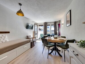 A compact room with a bed, a dining table with chairs, a TV mounted on the wall, and a window with curtains.