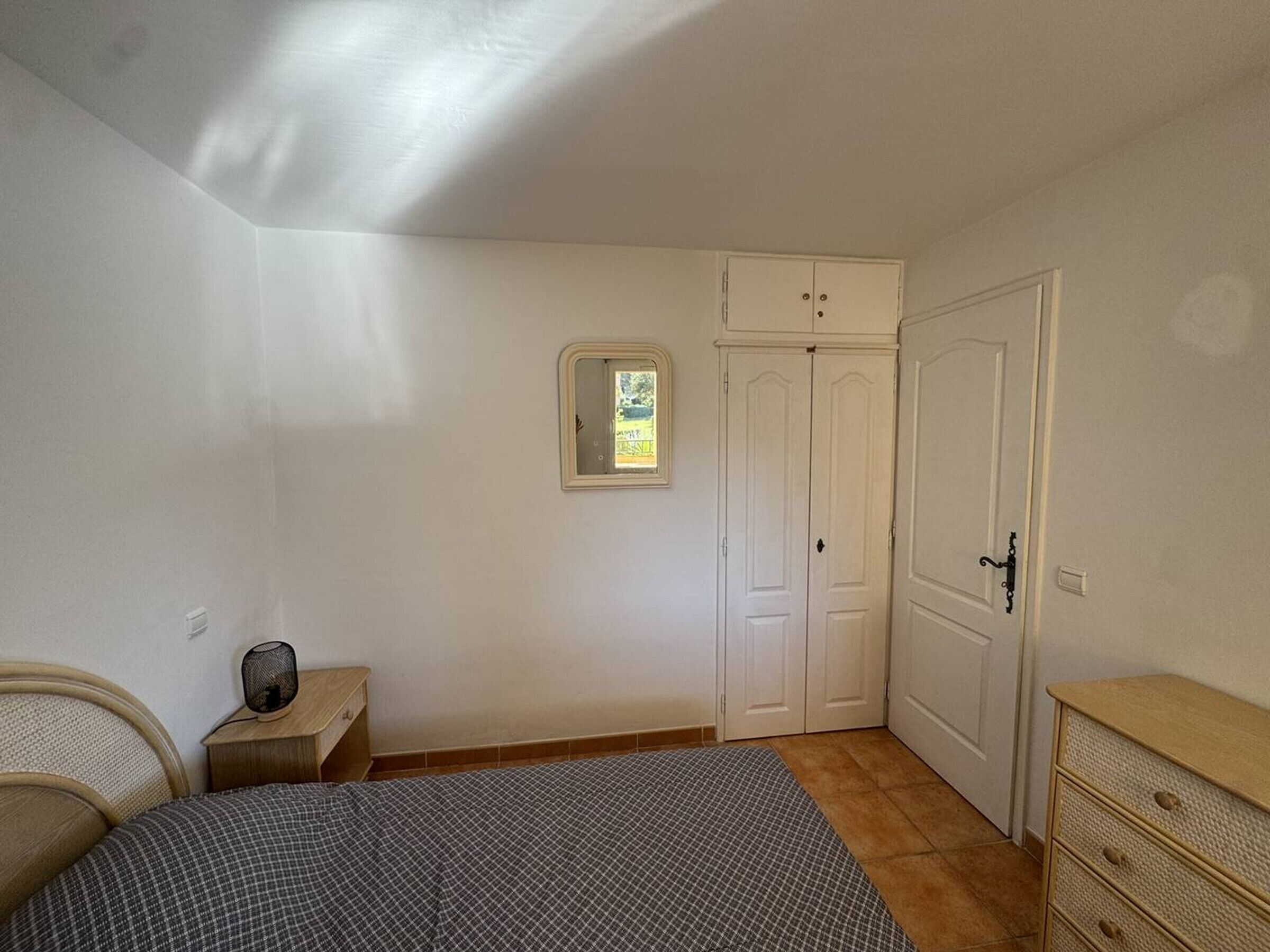 Een slaapkamer met een bed, nachtkastje met een ventilator, een dressoir en een kast.