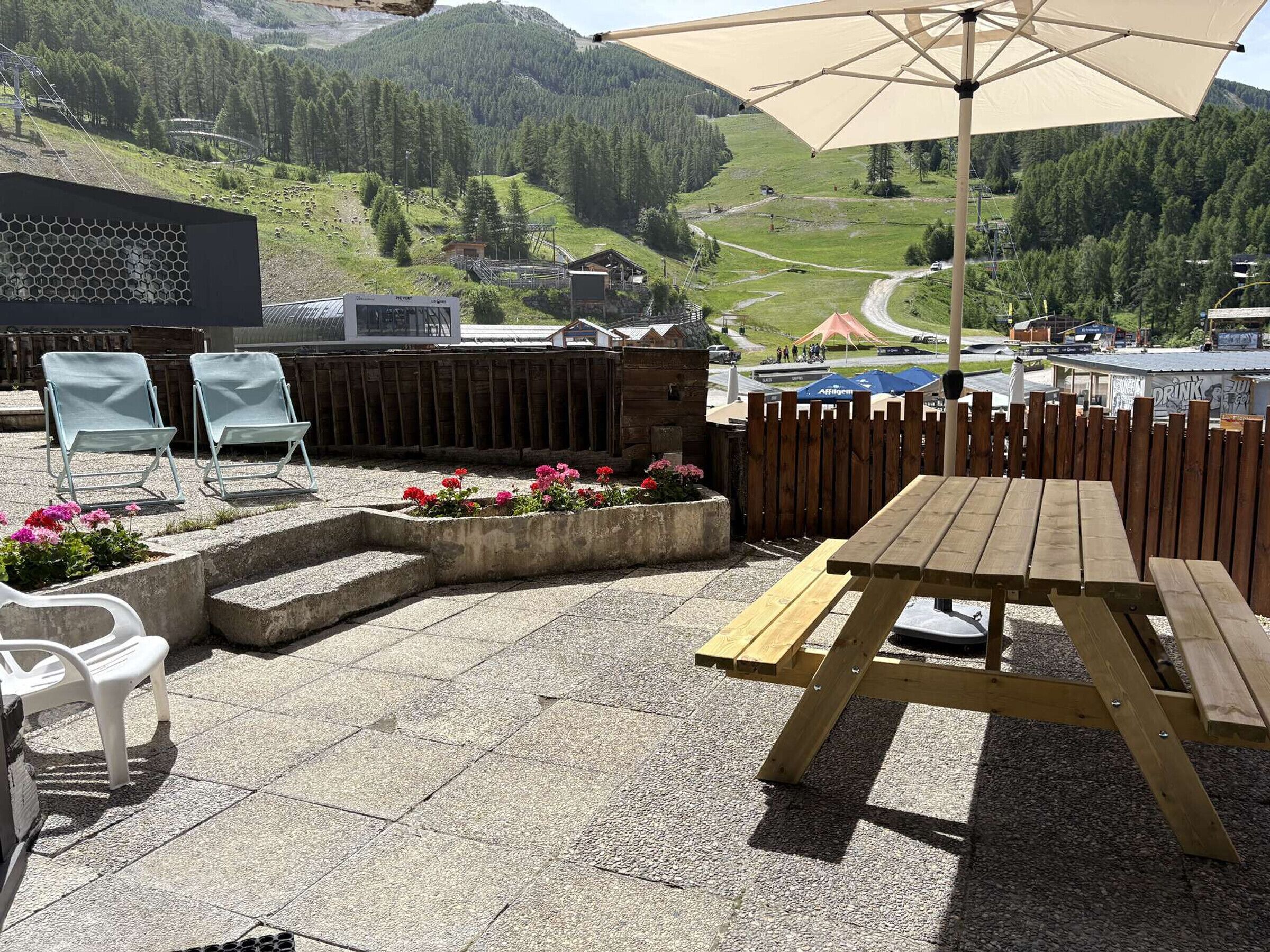 An outdoor patio with a wooden table and chairs, a concrete planter with flowers, and a view of a mountainous landscape.