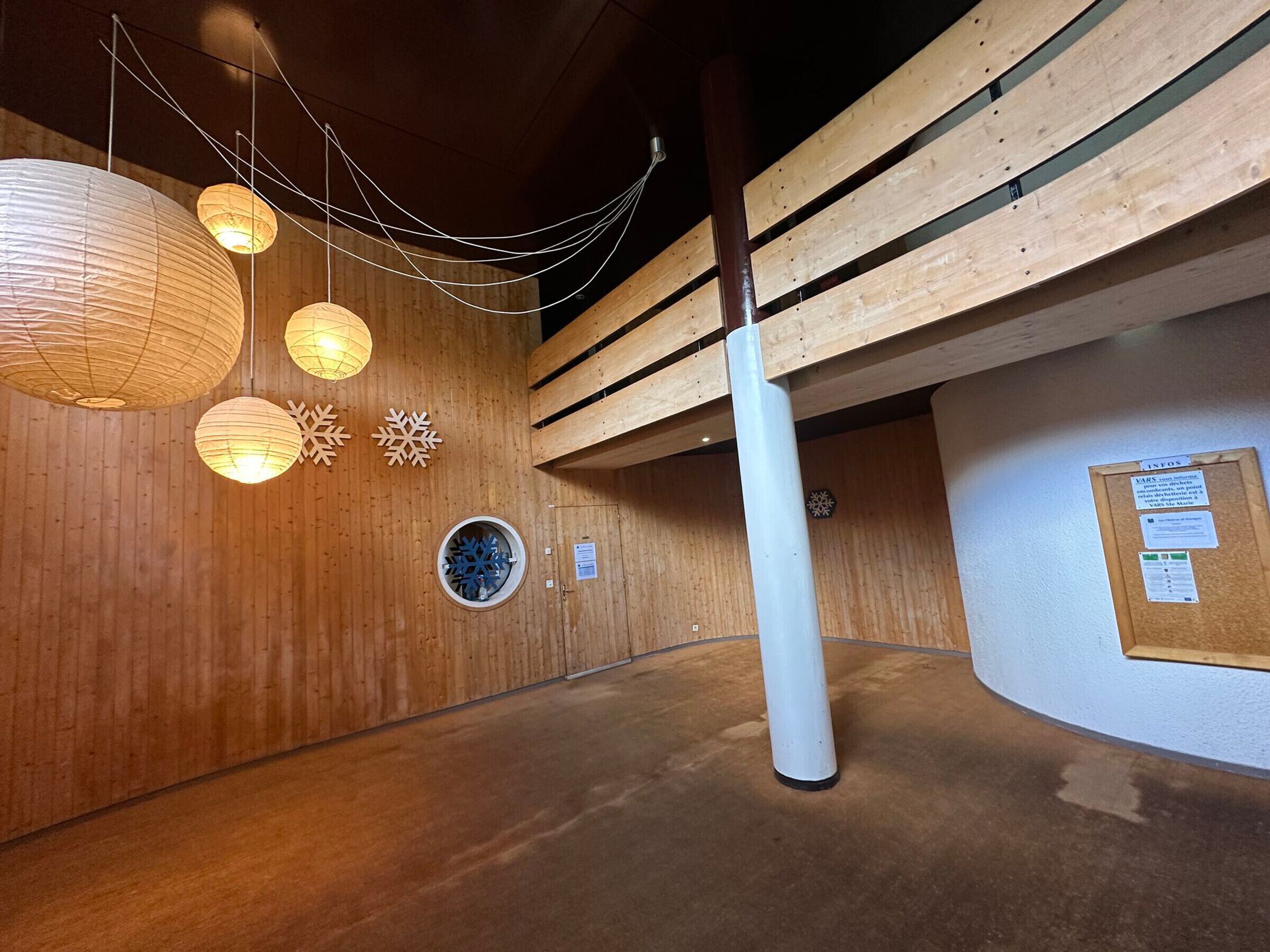 A wooden interior with hanging paper lanterns, a circular window with a snowflake design, and a bulletin board with notices.