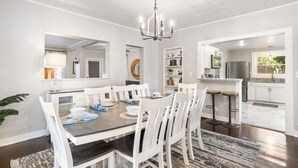 Un coin repas avec une table ronde, des chaises blanches et un lustre. On aperçoit une cuisine à l’arrière-plan, avec des armoires blanches et des appareils électroménagers en acier inoxydable.