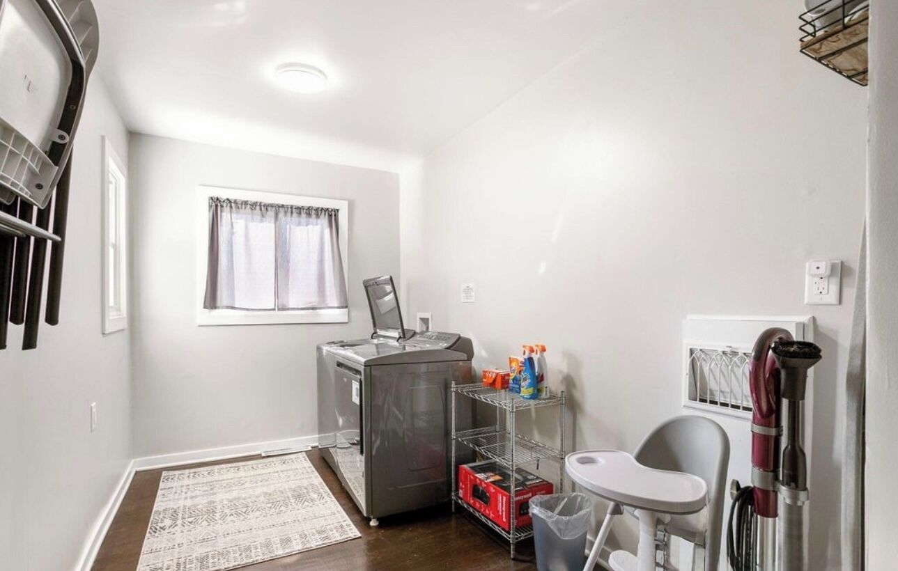 A compact kitchenette with a refrigerator, microwave, and small sink. There is a window with curtains, a rug, and a chair.