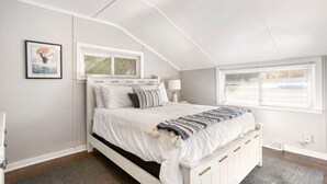 Une chambre avec un lit blanc, une fenêtre avec des stores, un tableau encadré au mur et un luminaire au plafond.
