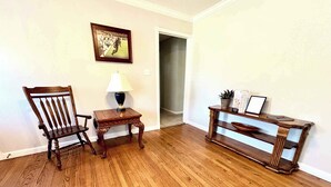 A room with hardwood flooring, a wooden chair, a small table with a lamp, and a console table with decorative items. - Retro Roost - 3BR/2.5BA - Close to Tech (Lubbock)