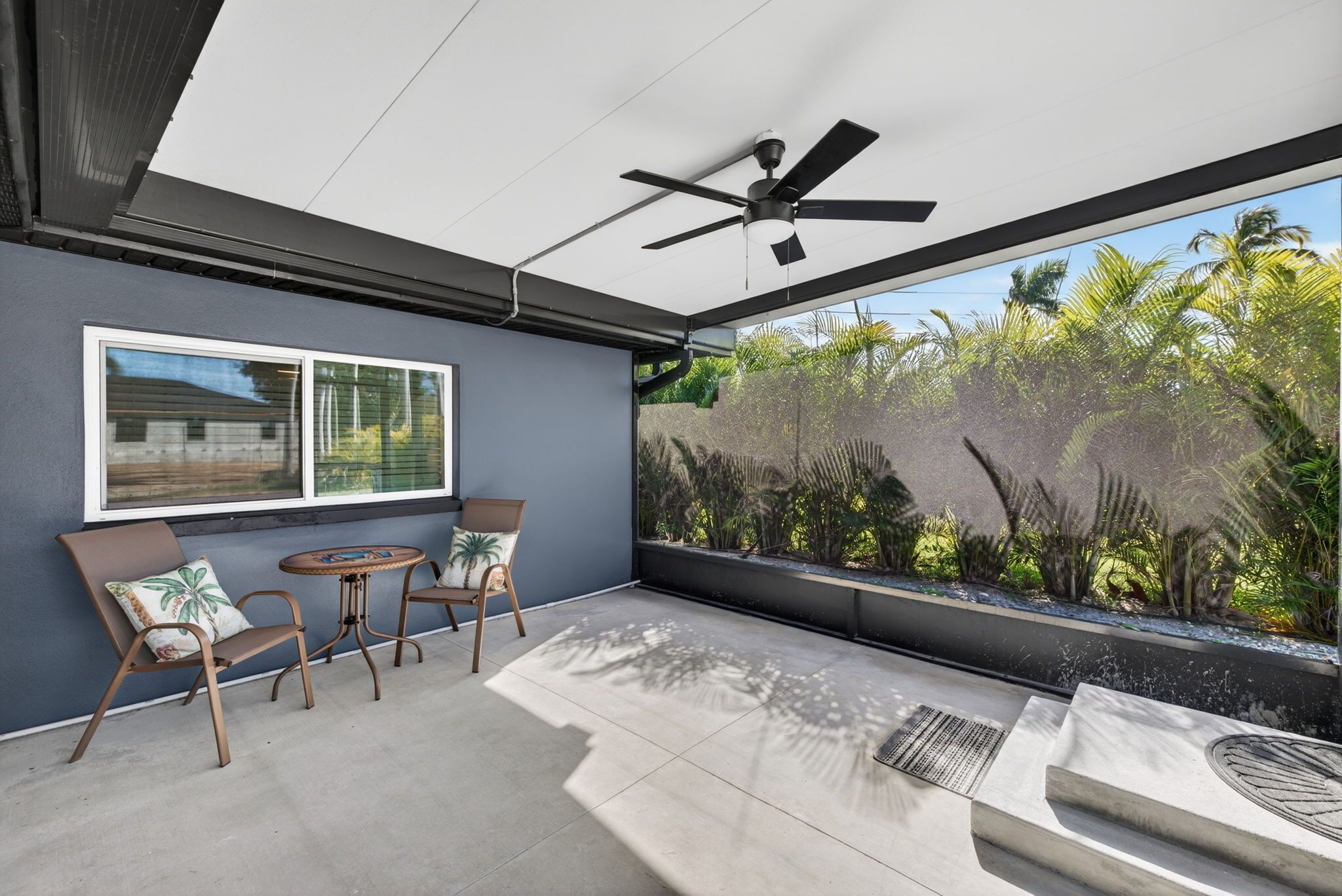 A modern patio with a ceiling fan, a window with blinds, a small round table, two chairs, and a view of tropical plants.