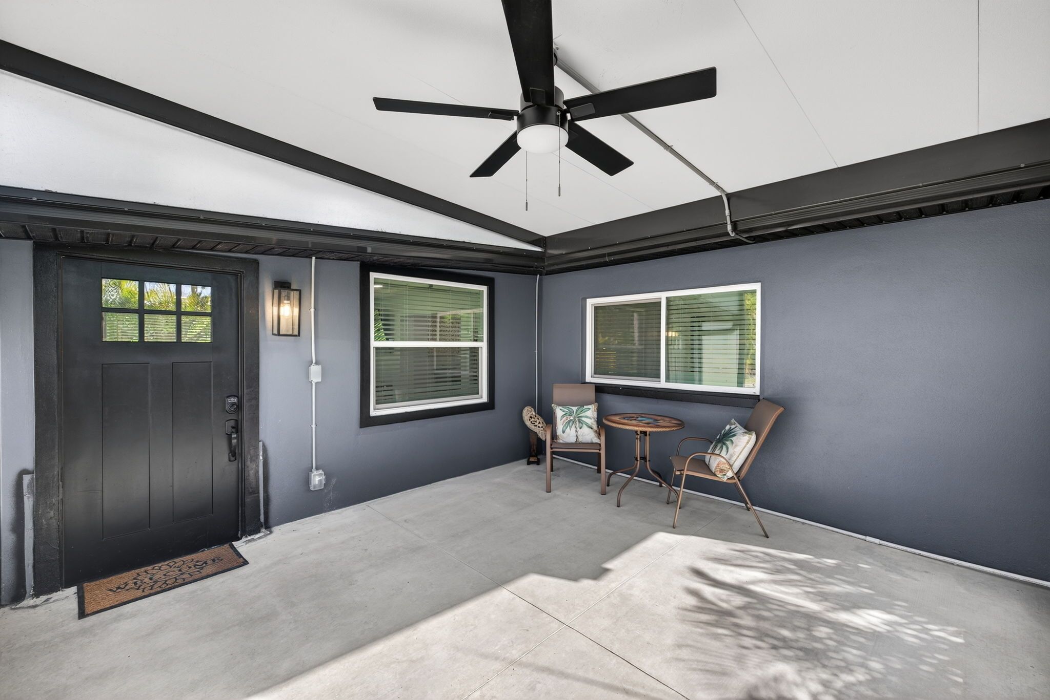 A room with a dark door, two windows with blinds, a ceiling fan, and a small round table with chairs.