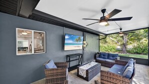 Un espacio de estar al aire libre cubierto con mobiliario de mimbre, un ventilador de techo y una pantalla de televisión mostrando una escena de playa.